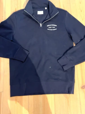 Mörkblå half-zip hoodie från Gant - Snygg mörkblå hoodie från Gant med halv dragkedja och hög krage. Tryck på bröstet med texten 'American Sportswear Gant 1949'. Tillverkad i mjukt bomullsmaterial och har ribbade muddar vid ärmar och nederkant. Perfekt för en avslappnad och stilren look.
