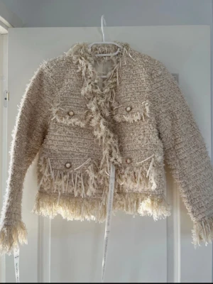 Beige bouclé cardigan med fransar - Trendig beige cardiganjacka i bouclé med fransiga detaljer längs kanter, ärmslut och nederkant. Jackan har två bröstfickor med dekorativa knappar och en öppen front. Perfekt för dig som gillar en chic och unik stil.