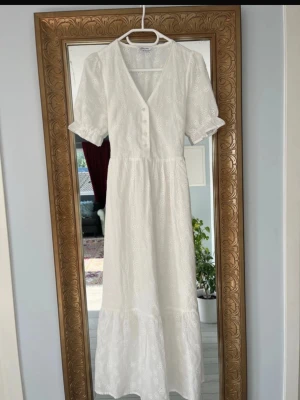Vit långklänning med broderade detaljer - Romantisk vit långklänning i bomull med broderade blommor, v-ringning och knappar framtill. Puffärmar med resår och knytband i midjan för en feminin siluett. Perfekt för sommarens alla tillfällen.