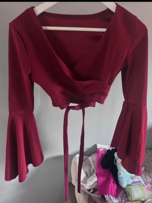 Vinröd omlottopp med trumpetärm - Snygg vinröd omlottopp med långa trumpetärmar och knytband i midjan. Toppen har en djup v-ringning och är croppad, vilket ger en trendig och ungdomlig look. Perfekt att styla med höga jeans eller kjol.