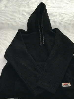 Svart fleecehoodie med huva - Mysig svart hoodie i teddyfleece med stor huva och dragsko. Hoodien har en magficka och en liten färgglad patch nertill. Perfekt för dig som gillar en avslappnad och varm stil.