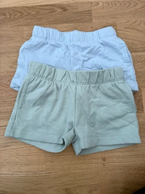 Två par mjuka shorts i pastell - Två par bekväma shorts i pastellfärger, ett par ljusgröna och ett par ljusblå. Båda har elastisk midja och är tillverkade i mjuk bomull, perfekta för varma dagar. Enkla och stilrena utan synliga detaljer eller mönster.