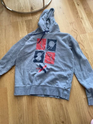 Grå hoodie med tryck 1983 - Snygg grå hoodie med huva och dragsko. På bröstet finns ett stort tryck med siffrorna 1983 i rött och mörkblått, samt detaljerna 428 och 306 under ett X. Hoodien har klassisk passform och ribbade muddar. Perfekt för en chill och avslappnad stil.