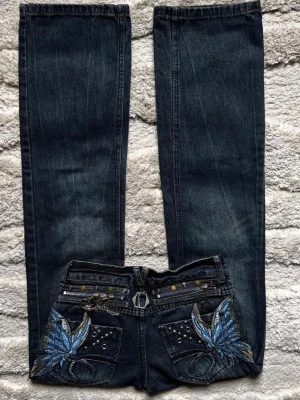 Mörkblå jeans med fjärilsbrodyr - Unika mörkblå jeans med raka ben och detaljerade broderier av blå fjärilar på bakfickorna. Jeansen har även nitar och dekorativa dragkedjor baktill för en extra cool look. Perfekta för dig som vill sticka ut med din stil.