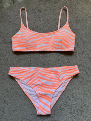 Vacker orangevit bikini med zebramönster - Snygg bikini i orange och vitt med zebramönster!😍SÅÅ COOL, Y2K vibe!! Toppen har smala justerbara axelband och rak skärning, samt spänne i ryggen. Bikinitrosan har klassisk passform. Perfekt set för stranden eller poolen i sommar! Toppen har vaddering som kan tas ut🤍Jätteskönt och tjockt material som INTE blir genomskinligt💞Använd några gånger, underdelen har inga fläckar. Hör av vid funderingar, pris diskuterbart❤️