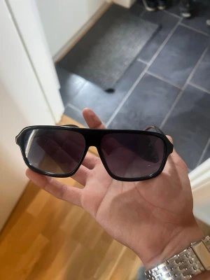 Svarta solglasögon från Hugo - Snygga svarta solglasögon från Hugo med bred båge och mörka, fyrkantiga glas. Klassisk och stilren design med diskret Hugo-logga på sidan. Perfekta för att lyfta din look under soliga dagar.