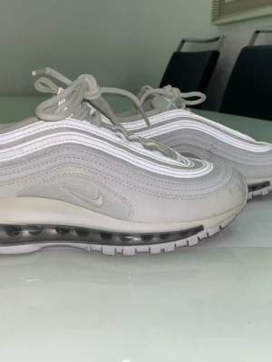 Nike Air Max 97 vita sneakers - Nike Air Max 97 sneakers i vitt och ljusgrått med ikoniska vågformade paneler och synlig Air-enhet i sulan. Ovandelen är i mesh och syntetmaterial, med reflekterande detaljer och klassisk snörning. Perfekta för dig som gillar streetwear och sportig stil.