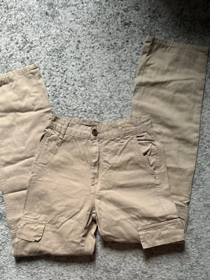 Beige cargobyxor med fickor - Snygga beige cargobyxor med raka ben och flera praktiska fickor, inklusive sidofickor med lock. Byxorna har bälteshällor och stängs med knapp och dragkedja. Tillverkade i ett mjukt bomullsmaterial som är skönt att bära.