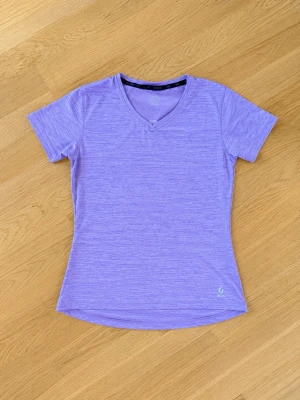 Lila träningströja från SOC - Snygg lila träningströja från SOC med V-ringning och korta ärmar. Tillverkad i ett lätt och snabbtorkande material som passar perfekt för löpning eller gym. Reflextape på ryggen för extra synlighet. Enkel och sportig design.