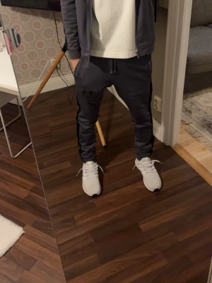 Mörkgrå träningsbyxor Under Armour  - Säljer ett par mörkgrå träningsbyxor från Under Armour med svarta detaljer längs sidorna och dragsko i midjan. Byxorna har en sportig passform och är perfekta för gymmet eller löpning. Materialet är mjukt och stretchigt för maximal komfort.