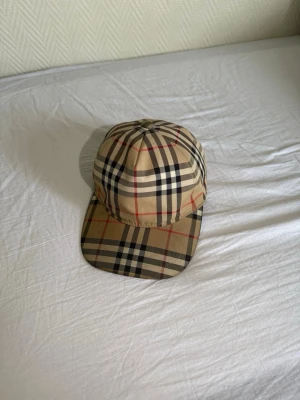  Burberry keps  - Säljer en klassisk beige keps från Burberry med det ikoniska rutiga mönstret i svart, vitt och rött. Kepsen har böjd skärm och justerbar snapback baktill. Märkt med 'BURBERRY LONDON ENGLAND' på baksidan. Perfekt för dig som vill ha en stilren och tidlös accessoar. Även lite använd men fortfarande i gott skick!