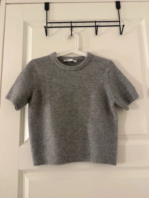 Grå stickad t-shirt  - Kortärmad stickad t-shirt i mjuk grå ullmix från Zara. Tröjan har rund halsringning och ribbade kanter vid ärmslut och nederkant. Perfekt att styla med jeans eller kjol för en chill look.