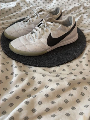 Vita Nike fotbollsskor med svart swoosh - Säljer ett par vita Nike fotbollsskor med svart swoosh på sidan. Skorna har en låg profil, snörning och slitstark ovandel i syntetmaterial. Sulan är ljusgrön och designad för bra grepp på inomhusplaner. Perfekta för dig som vill ha stilrena och funktionella skor till fotbollen.
