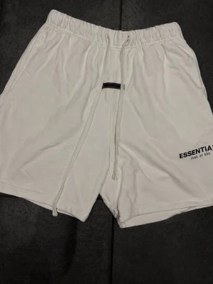 Vita shorts från Essentials Fear of God - Säljer ett par vita shorts från Essentials Fear of God med elastisk midja och snörning. De har en avslappnad passform och är perfekta för sommaren. Diskret logga på vänster ben och mjukt material för maximal komfort.