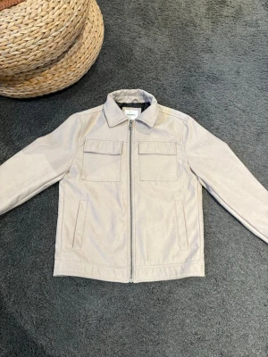 Beige overshirt med fickor från Dressman - Stilren beige overshirt/jacka från dressman med dragkedja framtill och två stora bröstfickor. Klassisk krage och raka ärmar ger en clean look. Perfekt att bära över en t-shirt för en avslappnad stil. Materialet ser ut att vara bomull eller bomullsblandning. Sitter bra på mig är 175 och väger 65. Endast använd en gång.