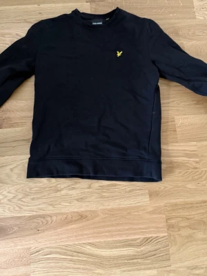 Svart sweatshirt från Lyle & Scott - Svart sweatshirt med rund hals från Lyle & Scott. Tröjan har en gul broderad logga på bröstet och är tillverkad i mjukt material. Perfekt för en avslappnad och stilren look. Priset ej hugget i sten. Hör av vid funderingar