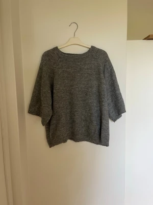 Grå stickad tröja - Mysig grå stickad tröja med rund halsringning och halvlånga ärmar. Tröjan har en enkel och stilren design, perfekt för lager på lager. Materialet är mjukt och bekvämt.