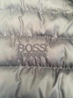 Svart dunväst från Hugo Boss - Snygg svart dunväst från Hugo Boss med diskret broderad logga på bröstet. Västen har ståkrage, dragkedja framtill och två praktiska fickor med dragkedja. Perfekt för lager-på-lager och ger en clean look till din outfit.