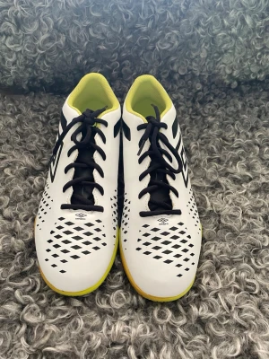 Vita Umbro fotbollsskor med svart mönster - Säljer ett par vita fotbollsskor från Umbro med svart mönster och svarta snören. Skorna har en gul insida och gummisula, samt ventilerande hålmönster på ovandelen. Perfekta för inomhusspel och ger bra grepp på planen.