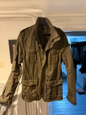 Olivgrön fieldjacket från H&M - Säljer en olivgrön fieldjacket från H&M med fyra stora fickor framtill och axelklaffar. Jackan har hög krage, knappar och dragkedja. Perfekt för dig som gillar en klassisk och avslappnad stil.