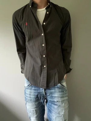 Svart skjorta från Ralph Lauren - Klassisk svart skjorta från Ralph Lauren med vit knappstängning och röd broderad logga på bröstet. Skjortan har lång ärm och normal passform, tillverkad i bomull. Perfekt för en stilren och avslappnad look.