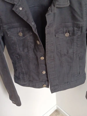 Svart jeansjacka från H&M - Snygg svart jeansjacka från H&M (&denim ) med klassisk krage, två bröstfickor med knapp och silverfärgade knappar framtill. Jackan har raka ärmar och en tidlös design som passar till det mesta. Perfekt för dig som vill ha en stilren och enkel jeansjacka!