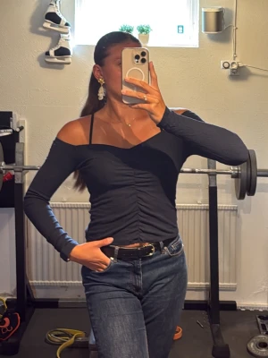 Märk blå offshoulder topp - Trendig svart offshoulder topp med långa ärmar och snygg rynkning framtill. Toppen har smala axelband och en figurnära passform som framhäver siluetten. Perfekt att styla med jeans eller kjol för en cool look.