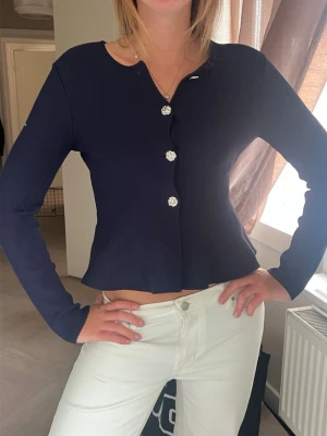Mörkblå cardigan med blommiga knappar - Snygg mörkblå cardigan med långa ärmar och dekorativa blommiga knappar framtill. Tröjan har en lätt croppad passform och rund halsringning. Perfekt att styla till både jeans och kjol för en trendig look.