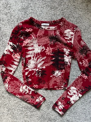 Röd croppad topp med drakmönster - Snygg röd croppad långärmad topp med asiatiskt inspirerat drakmönster i svart och vitt. Toppen har en tight passform och är tillverkad i ett stretchigt syntetmaterial. Perfekt för dig som vill sticka ut med en unik och trendig stil.