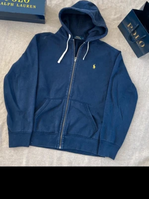 Mörkblå hoodie från Polo Ralph Lauren - Mörkblå hoodie med dragkedja från Polo Ralph Lauren. Klassisk design med vit dragsko i huvan, kängurufickor och den ikoniska gula loggan broderad på bröstet. Tillverkad i mjukt bomullsmaterial som är skönt att bära.