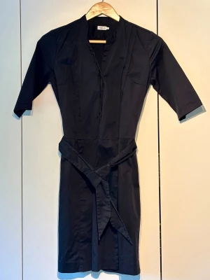 Svart klänning med bälte Filippa K - Stilren svart klänning från Filippa K med knytbälte i midjan och trekvartsärm. Klänningen har en rak passform och enkel design som ger en klassisk look. Perfekt för dig som gillar minimalistisk stil.