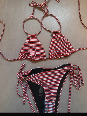 Randig bikini med knytband - Snygg bikini med röd, vit och blå randigt mönster. Överdelen har trekantsform och knytband i nacken och ryggen. Underdelen har matchande knyt i sidorna. Perfekt för stranden eller poolen.