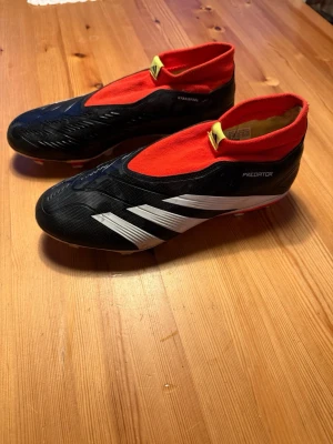 Adidas Predator fotbollsskor svart/röd - Säljer ett par Adidas Predator Pro fotbollsskor i svart med vita detaljer och röd strumpkrage. Skorna har en strömlinjeformad design utan snörning och är tillverkade i syntetmaterial. Perfekta för dig som vill ha bra grepp och kontroll på planen.