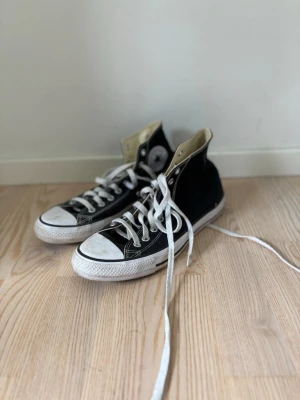 Svarta converse - Klassiska svarta höga sneakers med vita sulor och tåhättor. Skorna har vita snören och en rund logotyp på sidan. Ovandelen är i canvas och sulan i gummi. Perfekta för en avslappnad och cool stil.