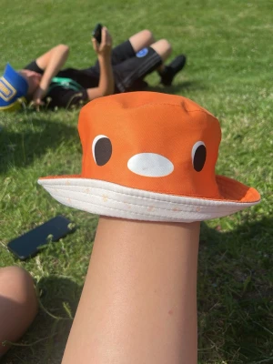 Orange bucket hat med fiskmotiv LIMTED EDITION.KAN FÖRHANDLAS  - Säljer en orange bucket hat med vitt brätte och ett gulligt fiskansikte framtill. Hatten är i mjukt tyg, troligen bomull, och har svarta och vita detaljer som ögon och mun. Perfekt för soliga dagar och ger en lekfull vibe till din outfit. Kan förhandlas 