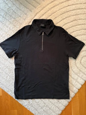 Svart piké-tröja med zip dragkedja - Säljer en svart piké från Selected Homme med korta ärmar och en snygg dragkedja framtill. Toppen har klassisk krage och är gjord i mjukt bomullsmaterial. Perfekt för en clean och stilren look. Fint skick!
