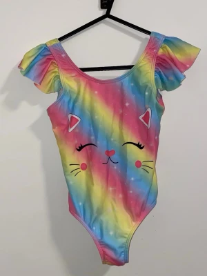 Regnbågsfärgad baddräkt med kattmotiv - Söt baddräkt i regnbågsfärgat tyg med volangärmar och tryckt kattansikte framtill. Färgerna går i rosa, blått, gult och grönt med små vita prickar. Rundad halsringning och låg rygg. Perfekt för strand eller pool!
