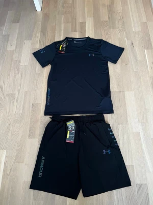 Svart träningsset från Under Armour - Snyggt svart träningsset från Under Armour med t-shirt och shorts. T-shirten har korta ärmar och diskret logga på bröstet. Shortsen har elastisk midja, logga på benet och är tillverkade i ett lätt, snabbtorkande material. Perfekt för gym eller löpning.