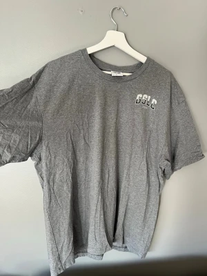 Grå Gymshark t-shirt - Snygg grå t-shirt från Gymshark i storlek XL med GSLC-tryck på bröstet. Klassisk rund hals och korta ärmar. Tillverkad i mjuk bomull som passar perfekt till en avslappnad stil.