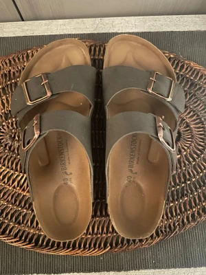 Bruna Birkenstock sandaler med spännen - Säljer ett par klassiska bruna Birkenstock sandaler med två breda remmar och justerbara metallspännen. Fotbädden är ergonomiskt utformad för extra komfort och sulan är i kork. Perfekta för en avslappnad och stilren look.