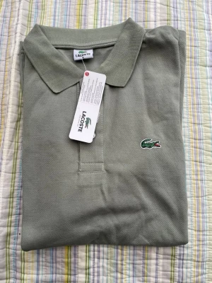 Grön pikétröja från Lacoste - Snygg grön pikétröja från Lacoste med klassisk krage och knappslå framtill. Tröjan har den ikoniska krokodilloggan broderad på bröstet och är tillverkad i ett svalt bomullsmaterial. Perfekt till jeans eller shorts.