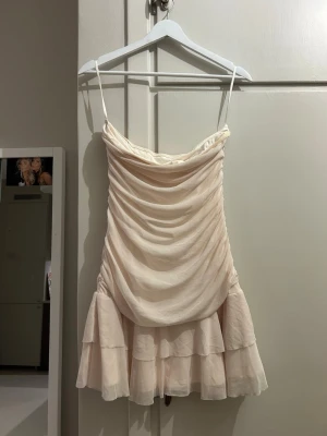 Ljus kort klänning med volang - Söt ljus kort klänning med tunna axelband och draperad framsida. Klänningen har volangdetaljer nedtill och är perfekt för sommarens varma dagar. Tillverkad i ett tunt, mjukt material som ger ett romantiskt intryck.