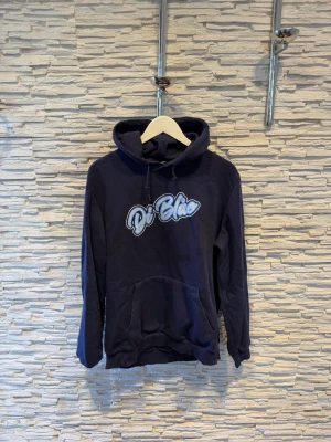 Mörkblå hoodie med tryck ”Di Blåe” - Snygg mörkblå hoodie med stor magficka och huva med dragsko. På bröstet finns ett vitt och blått tryck med texten 'Di Blåe'. Hoodien har ribbade muddar vid ärmar och nederkant. Perfekt för en avslappnad och cool stil.
