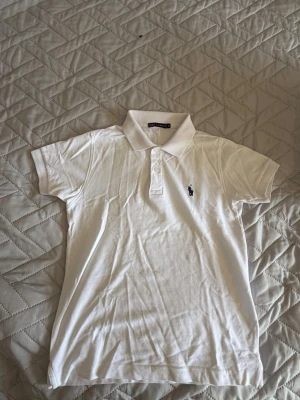 Vit pikétröja från Ralph Lauren - Klassisk vit pikétröja från Ralph Lauren med korta ärmar och den ikoniska broderade loggan på bröstet. Tröjan har krage och knappar framtill. Tillverkad i mjuk bomull som känns skön mot huden. Perfekt till jeans eller shorts för en clean look.