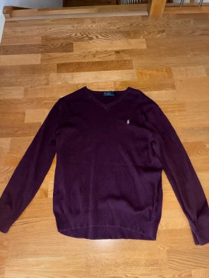 Vinröd stickad tröja Polo Ralph Lauren - Snygg vinröd stickad tröja från Polo Ralph Lauren med V-ringning och logga broderad på bröstet. Tröjan har långa ärmar och ribbade muddar vid ärmslut och nederkant. Perfekt för lager på lager och stilren look.