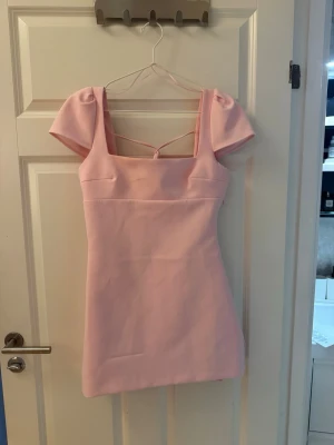 Rosa kort klänning med puffärm - Söt ljusrosa kort klänning med puffärmar och snygga knytband i ryggen. Klänningen har fyrkantig urringning och är figurnära i modellen. Perfekt för dig som vill sticka ut med en feminin och trendig look.