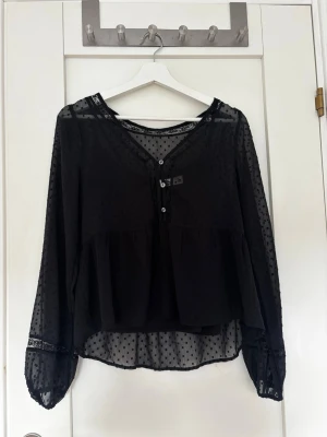 Svart transparent blus med prickar - Snygg svart blus i transparent material med små sammetsprickar och volang nedtill. Blusen har v-ringning med knappar framtill och långa ärmar med spetsdetaljer och knapp vid ärmslut. Den har även ett inbyggt linne. Perfekt för en trendig och chic look.