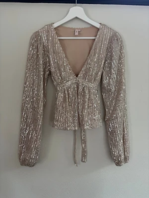 Glittrig beige topp från Nelly - Supersnygg glittrig topp från Nelly i beige med paljetter över hela. Djup v-ringning, långa ärmar och knyt i midjan ger en trendig look. Perfekt för dig som vill sticka ut och glänsa lite extra. Materialet känns mjukt och följsamt.