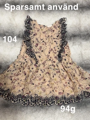Blommig beige volangklänning - Söt beige klänning med blommigt mönster och volangdetaljer i mörkblått längs axlar och nederkant. Klänningen har en luftig kjol och är ärmlös, perfekt för varma dagar. 🌸Sparsamt använd. Storlek 104