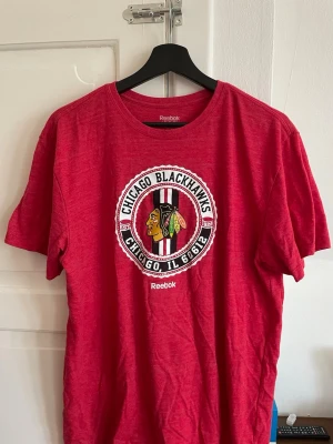Röd Chicago Blackhawks t-shirt Reebok - Säljer en röd t-shirt från Reebok med Chicago Blackhawks-logga tryckt framtill. T-shirten har rund halsringning och korta ärmar. Perfekt för dig som gillar hockey och vill ha en sportig stil.
